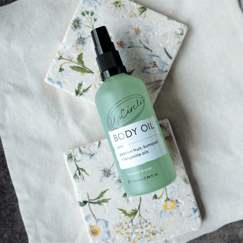 A bottle of UpCircle body oil with passion fruit, kumquat, and tangerine oils, placed on a floral patterned surface.