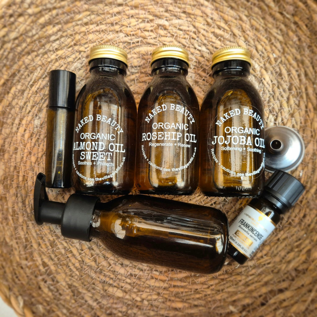 A set of handmade cosmetic and fragrance oil bottles arranged on a woven straw mat. The collection includes three amber glass bottles with gold caps labeled 'NAKED BEAUTY Organic Almond Oil', 'Organic Rosehip Oil', and 'Organic Jojoba Oil', a small black spray bottle, a black pump bottle, a silver metal cap, and a small essential oil bottle labeled 'Frankincense'.
