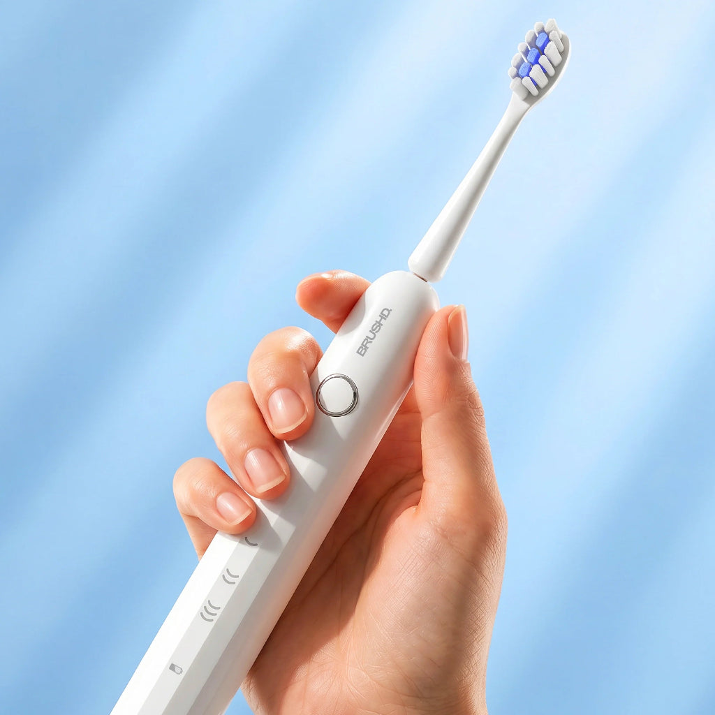 Hand holding a white electric toothbrush with a blue bristle head against a light blue background