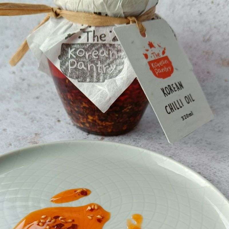 Glass jar of Gochugaru Chilli Oil VEGAN (250ml) with a dish of vibrant vegan chili oil and seeds.