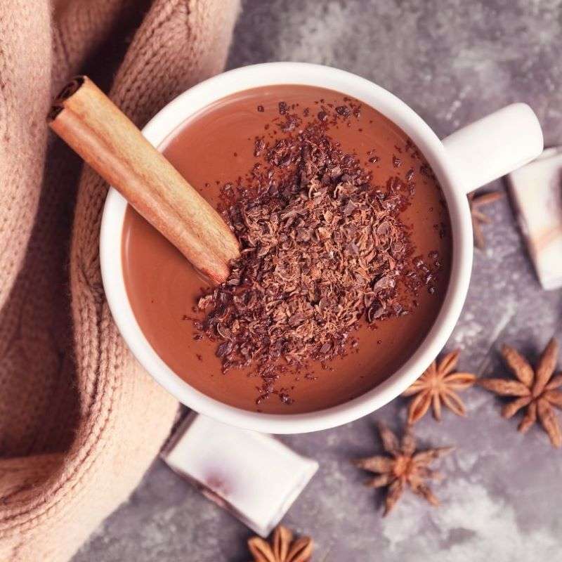 Cup of Drinking Chocolate Flakes (per 100g) topped with cinnamon stick, surrounded by spices and cozy fabric on table