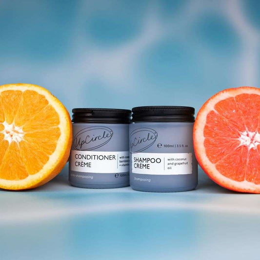Two containers of UpCircle hair care products with half an orange and half a grapefruit on a blue background