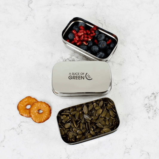 Small metal snack tin with seeds and dried fruit on a white marble surface, branded 'A Slice of Green'.