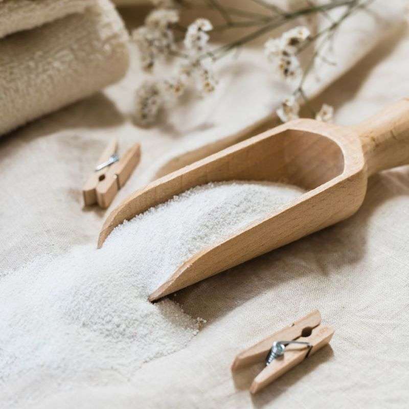 Wooden scoop filled with white powder on a beige surface with small wooden clothespins and dried flowers.