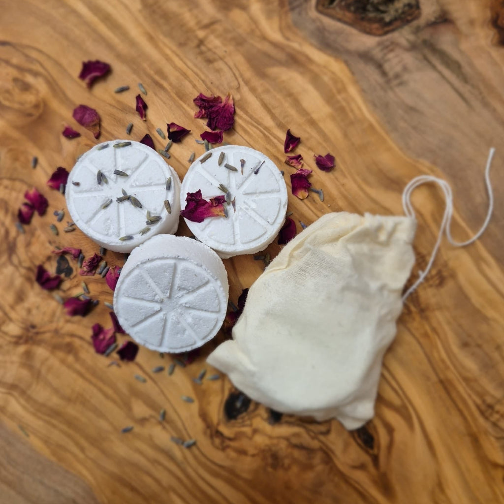 Three white shower steamers with dried lavender and rose petals on an olive wood board, with a small muslin drawstring bag