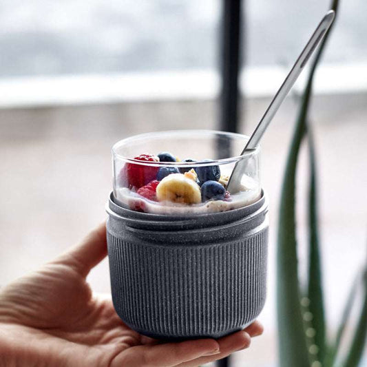Hand holding a glass of yogurt with berries and bananas, wrapped in a gray sleeve, with a blurred indoor background.