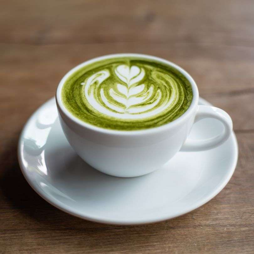Cup of creamy latte made with Matcha Green Tea ORGANIC (per 100g) in a white ceramic cup on wooden table