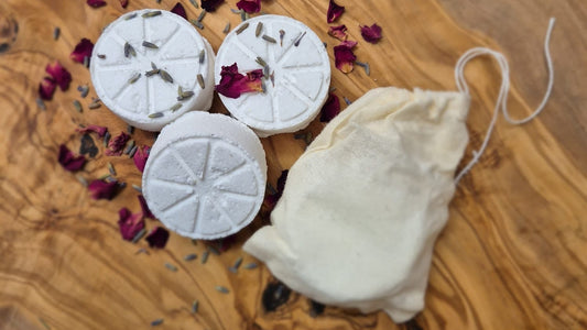 Three white shower steamers with dried lavender and rose petals on an olive wood board, with a small muslin drawstring bag