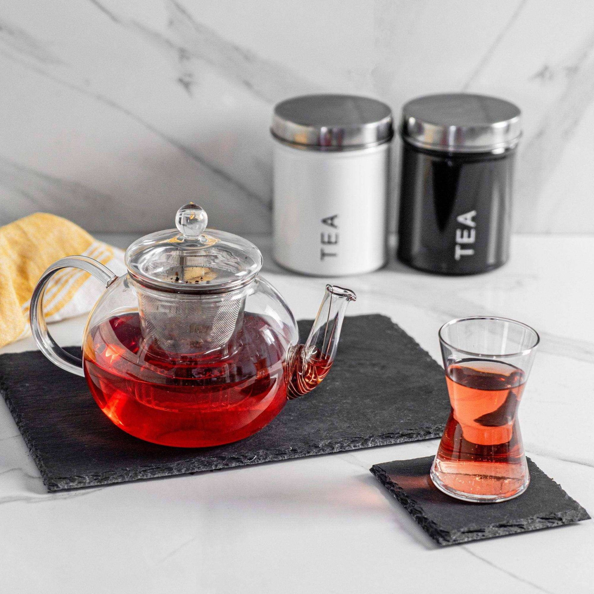 Clear glass teapot with red liquid on a black coaster, accompanied by a glass of tea and two metal tea canisters.