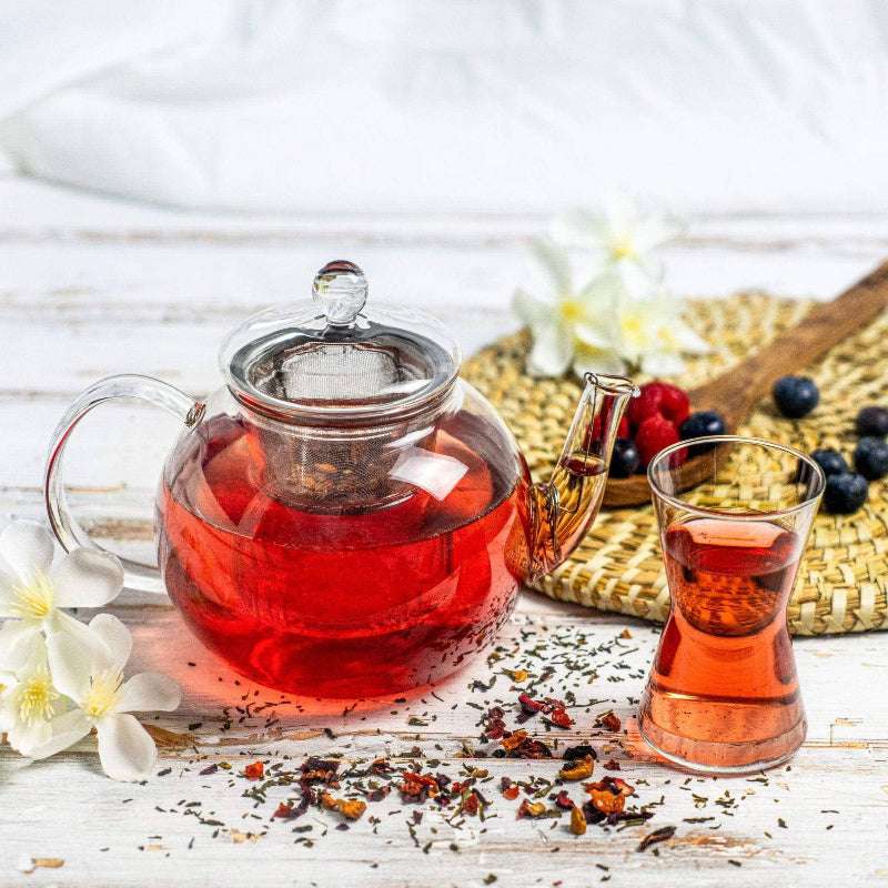 Glass teapot and cup with red tea on a wooden surface with flowers and berries.