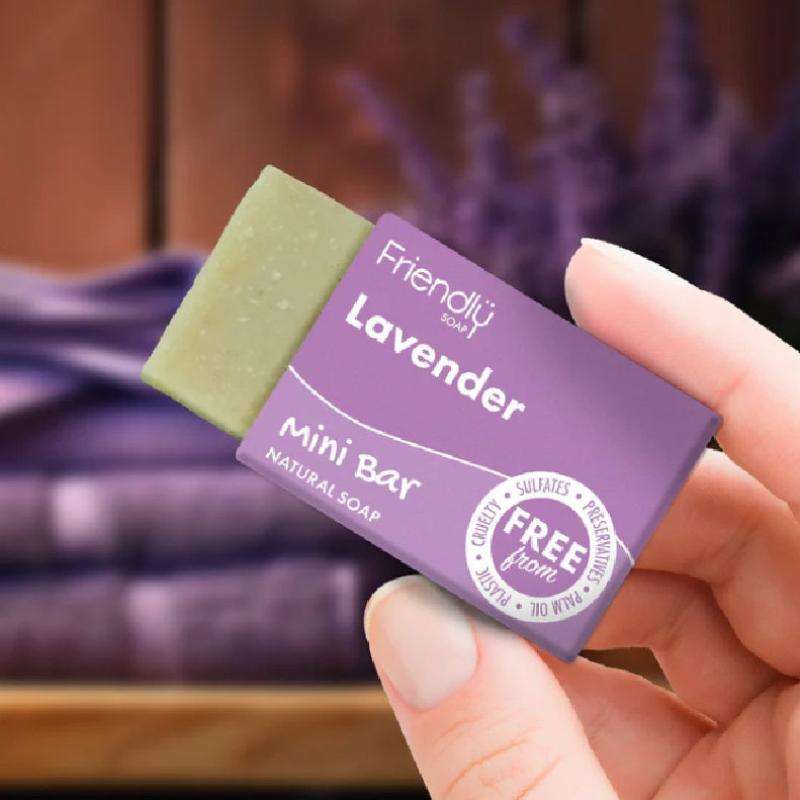 Hand holding a lavender mini bar soap with a blurred background of lavender flowers.