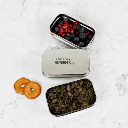 Small metal snack tin with seeds and dried fruit on a white marble surface, branded 'A Slice of Green'.