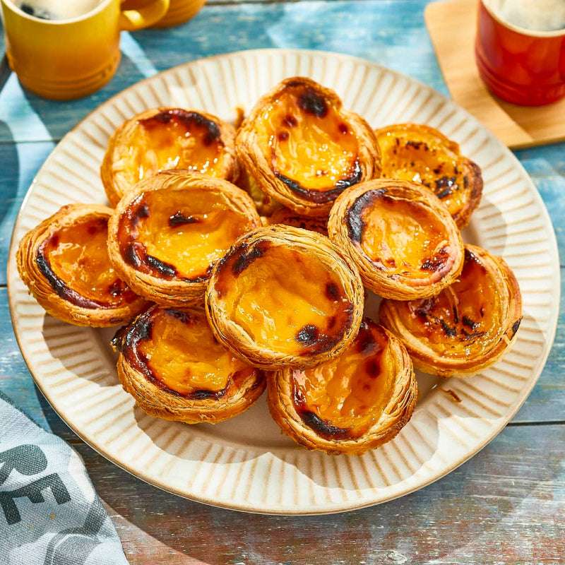 Plate of Portuguese egg tarts on a wooden table with mugs in the background
