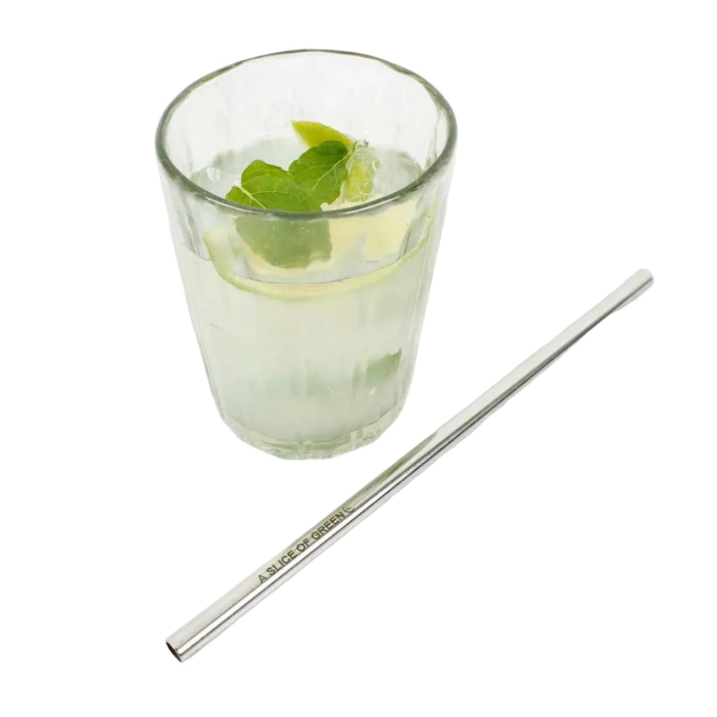 A stainless steel metal straw with 'A. Slice of Green' printed on it, placed on a white surface next to a glass of iced drink with mint and lime.