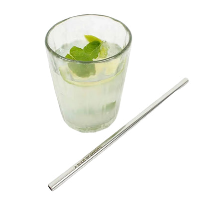 A stainless steel metal straw with 'A. Slice of Green' printed on it, placed on a white surface next to a glass of iced drink with mint and lime.