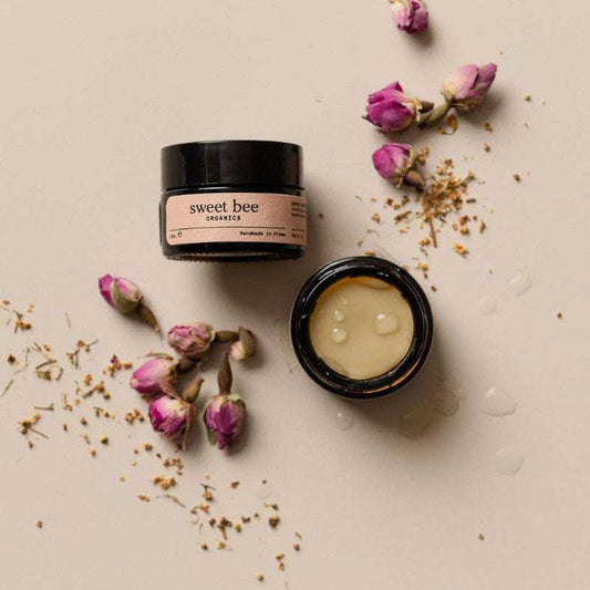 Two black jars with 'sweet bee' branding on a beige surface with dried flowers and petals.