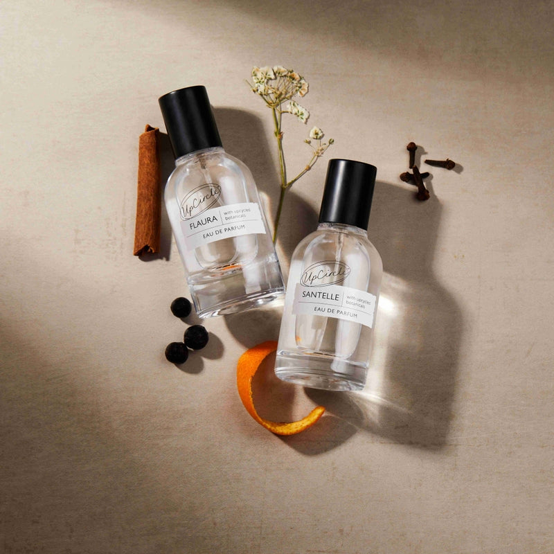 Two perfume bottles labeled 'Raura' and 'Santelle' on a textured surface with a cinnamon stick, orange peel, and blackberries.
