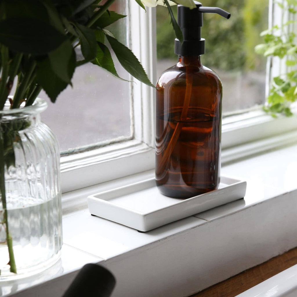 An amber glass pump bottle with a black dispenser sits on a white tray on a windowsill, with a glass vase of flowers nearby and natural light coming from outside.