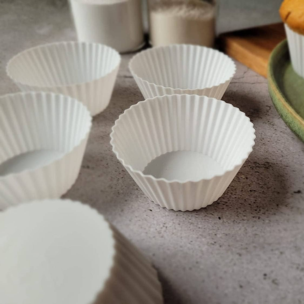 White fluted silicone cupcake liners arranged on a gray countertop, with baked chocolate chip muffins in paper liners on a green plate in the background.