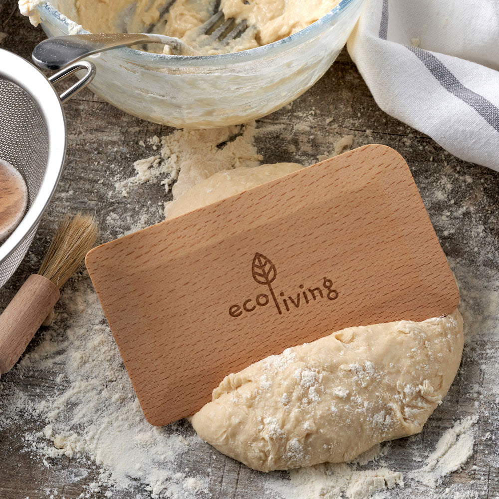 A wooden dough scraper with the brand name 'ecoliving' engraved on it, resting on flour-dusted dough on a rustic wooden surface with baking tools in the background.