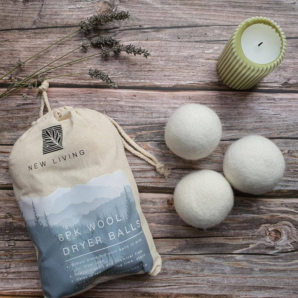 A set of six wool dryer balls by New Living, shown with their beige drawstring storage bag on a rustic wooden surface, accompanied by a green candle and dried lavender sprigs.