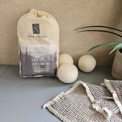A beige drawstring pouch labeled 'New Living 6pk Wool Dryer Balls' stands on a gray surface next to three cream-colored wool dryer balls, with a folded woven textile and a potted plant in the background.