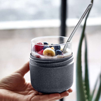 Hand holding a glass of yogurt with berries and bananas, wrapped in a gray sleeve, with a blurred indoor background.
