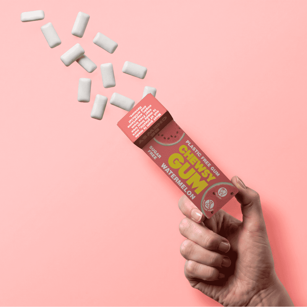 A hand holds an open tube of gum against a solid pink background, with white gum pieces spilling out in a diagonal line. The tube is red with yellow and green text and watermelon graphics.