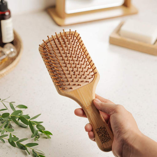Hand holding a bamboo hairbrush on a light surface with a plant and bottle in the background