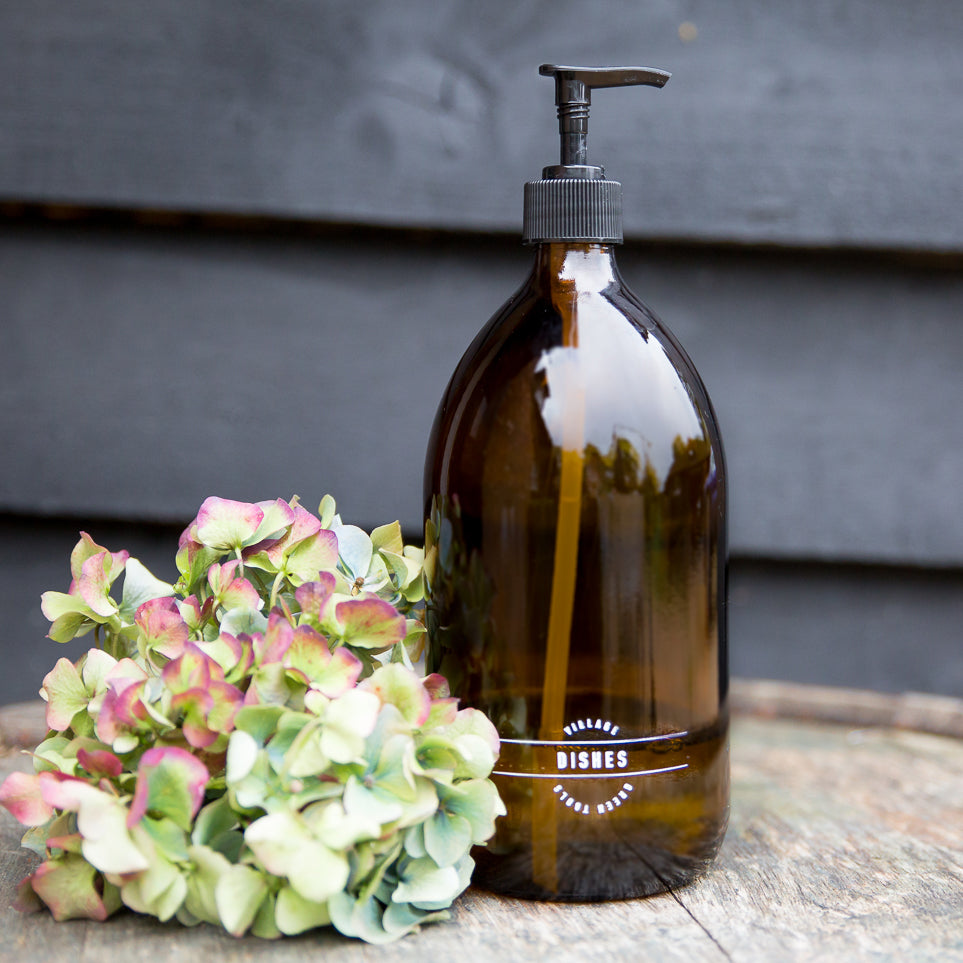 A brown glass refillable dish soap bottle with a black pump, placed next to some flowers.