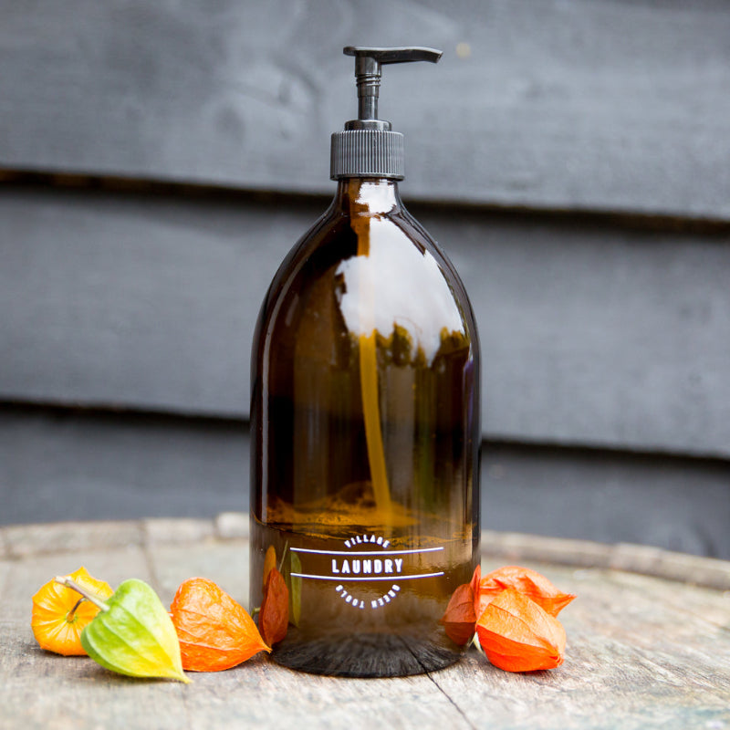 A clear glass amber refillable bottle with a black pump, labeled 'LAUNDRY', filled with a liquid, placed on a wooden surface with some orange peel slices and leaves around it.