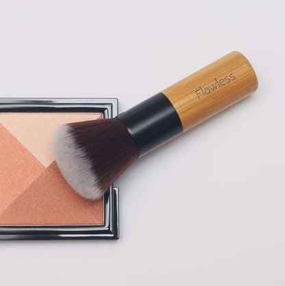 Makeup brush with wooden handle labeled 'Flawless' next to a compact powder on a light gray background