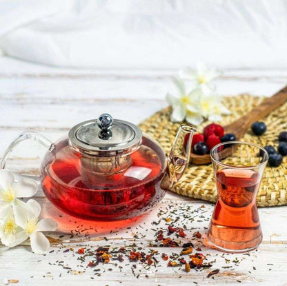 Tea set with a teapot and glass containing red tea, surrounded by dried herbs and berries on a wooden surface.